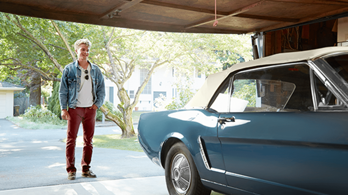 Middle aged man with red pants and a jean jacket, sun shining in the background. Standing at the opening of a garage, looking lovingly at a light blue with cream soft top antique vehicle. 